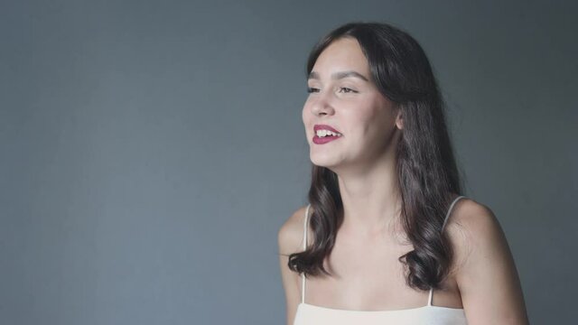 portrait happy laughing caucasian young female brunette woman mocking and giggling with mouth opened looking right on grey background. student white naked shoulders red lips long hair touching fringe