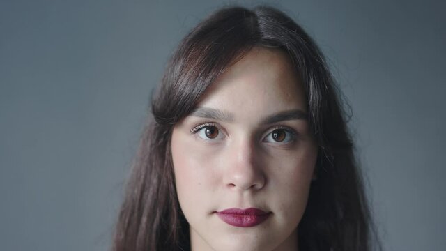 closeup portrait of pretty young brunette caucasian female looking at camera with big brown eyes and dark red lipstick on a grey background. emotions and facial view. long straight hair with fringe