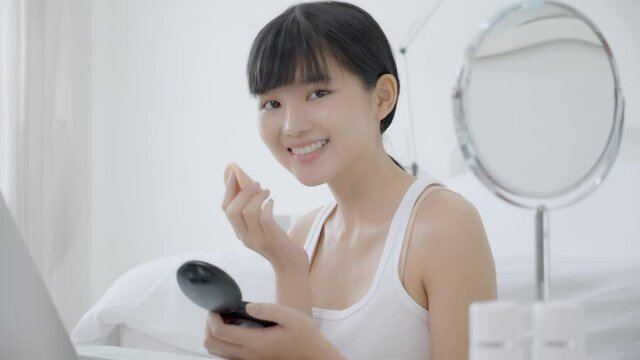 Beauty of young asian woman with learning makeup with powder puff on cheek watching laptop computer with tutorial course online, female teaching and explain make up with cosmetic on streaming.