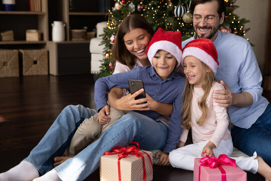 Happy bonding young couple parents and laughing little kids using cellphone, posing for funny selfie web camera photo, holding video call conversation, sitting on floor near decorated Christmas tree.