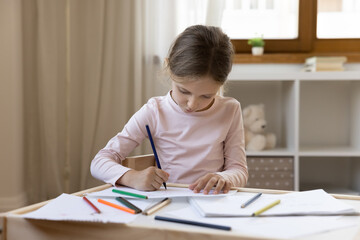 Focused adorable small preteen child girl drawing pictures with pencils on paper enjoying creative hobby activity, developing art skills sitting at table alone at home, art skills development.