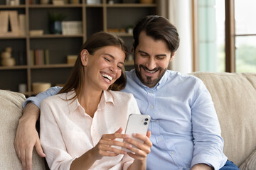 Happy laughing young family couple using funny mobile apps, watching funny photo video content in social network, entertaining sitting on cozy sofa at home, modern tech gadgets addiction concept.
