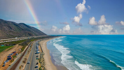  an aerial shot of the vast blue ocean water, lush green farmland, cars on the road, mountain ranges, thick cloud cover with blue sky and a rainbow at Rincon Beach in Ventura County California