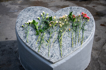 A large heart shaped grey granite blank headstone. The monument has small black flecks in it. The top and sides are smooth. There are multiple long stemmed carnations both white and pink on top. 