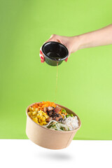 A woman's hand pours soy sauce into a floating bowl of salad poke and tuna, carrot, cucumber, and pineapple with a green background.