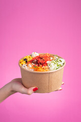 a pink bottom salad poke held by a woman hand with red nails, hawaiian asian food, chopsticks, carrot, green beans, pineapple, soy, jicama, seaweed, healthy food