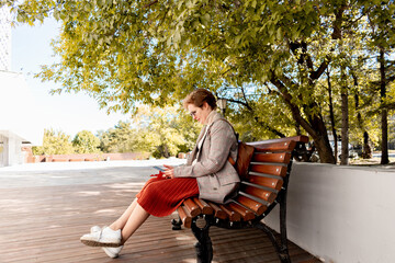 Fototapeta premium Woman with short red hair sits on a bench in the city. People browses social networks.
