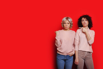 Portrait of two sad young women standing together and looking at camera and points at copy space isolated over red background