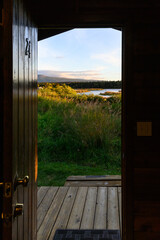 View out the door of a rustic wood cabin of a sunny landscape with tall grasses, a river, and blue sky
