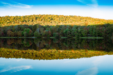 Waneta Lake, Catskills, New York State, USA - Forested hill at edge of calm lake, leaves beginning to change in Autumn and reflection of trees and shadows in water.  Blue sky with wispy clouds. 