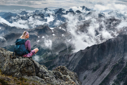 Adventurous woman sitting on cliff over mountains and clouds relaxing and meditating. Whistler. Garibaldi Provincial Park. British Columbia. Canada