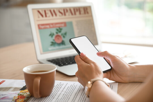 Young woman using mobile phone on table with cup of coffee, newspaper and laptop