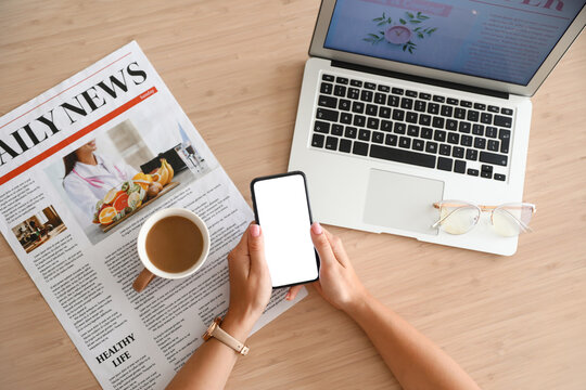 Female hands with mobile phone, newspaper, laptop and cup of coffee on wooden table