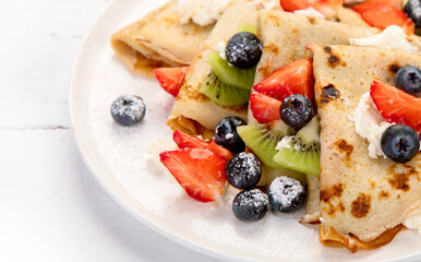 Sweet crepes with berries and cream on white wooden background.