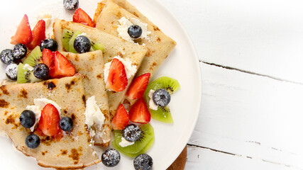 Sweet crepes with berries and cream on white wooden background.