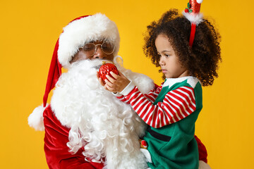 Little African-American girl with Santa Claus and Christmas ball on yellow background