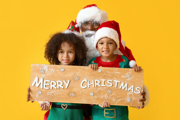 Little elves and Santa Claus holding board with text MERRY CHRISTMAS on yellow background - obrazy, fototapety, plakaty