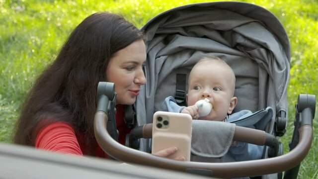 Mother and newborn baby doing selfie in park, mom with six month old baby boy in infant kid carriage on a walk outdoors. High quality 4k footage