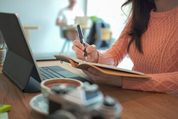 Young female in pink sweater using computer table and making notes on notebook.