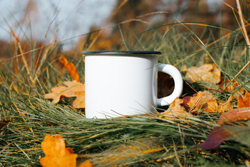 Camping enameled white mug mock-up standing on green grass with fallen autumn leaves, outdoors....
