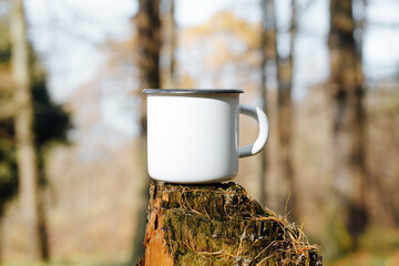 Camping metal white mug mock up standing on tree stump in woods outdoors. Enamelled balnk cup with...