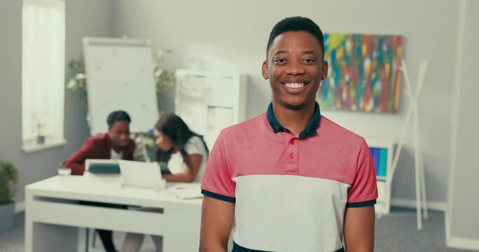 Portrait of young man with dark eyes hair black skin beautiful wide white smile man is wearing salmon collar shirt student in classroom is looking at window then at camera