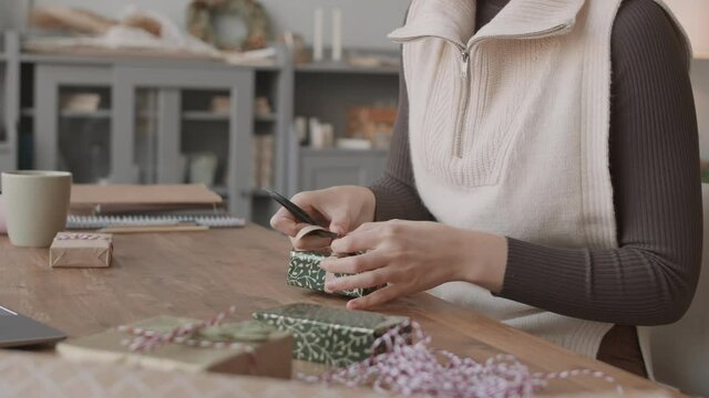 Locked-down of cropped unrecognizable woman sitting at desk in craft shop, frizzing ribbons on little gift boxes at daytime