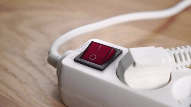 Finger is switching off a white electrical extension cord, pushing button close up. Saving electricity concept. Red switch. Connected electric plug. On a wooden background