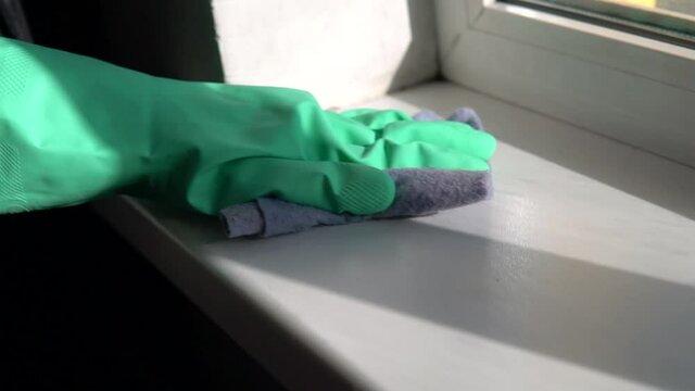 Woman hand palm in a rubber glove is wiping white plastic windowsill surface with wet cloth. Home apartment cleaning work in a sunny day.