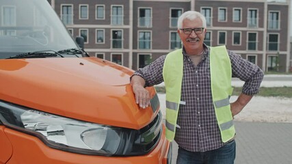 Medium handheld portrait of friendly senior bus driver in acid green vest smiling to camera standing outdoors leaning on orange bonnet of modern passenger bus