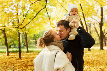 Fototapeta premium Happy young parents with a small child are kissing in a beautiful autumn park. Love, tenderness and relationships.