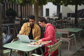Young man with Down syndrome with his mentoring friend sitting outdoors in cafe using laptop.