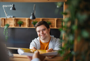 Happy young man with Down syndrome sitting and studying indoors at school, taking book from mentor - obrazy, fototapety, plakaty