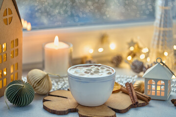 Fototapeta na wymiar Chocolate drink with marshmallow, cinnamon and milk froth. Christmas seasonal beverage on decorated windowsill. Wintertime. Candles and garland blurred background, selective focus