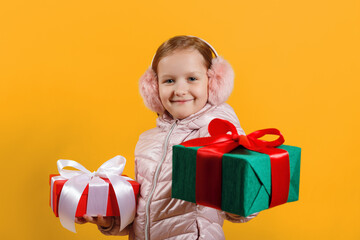 Fototapeta na wymiar Happy little girl holds out a box with a gift. Child in a jacket and warm headphones on a yellow isolated background. Christmas new year winter concept