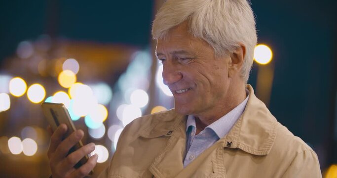 Bokeh shot of senior smiling man using smartphone on city street at night