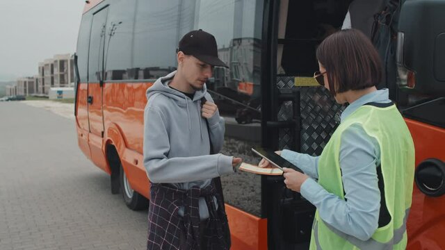 Medium handheld shot of teenage girl and young man getting on bus, showing electronic or printed tickets to female bus controller in acid green vest scanning them on digital tablet