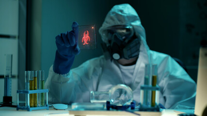 Scientist in ppe suit looking at small transparent tablet with biohazard hologram in lab