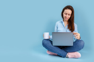 A young adult girl is sitting with a laptop on her lap and holding a white mug in her hands while looking into a laptop. Blue background with copyspace