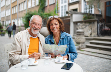 Fototapeta na wymiar Portrait of happy senior couple tourists sitting and using map outdoors in town, looking at camera.