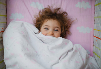 Top view of happy little girl lying in bed hiding under blanket, looking at camera indoors at home