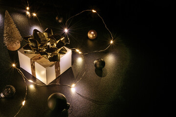 Christmas gift. White gift with golden bow, gold balls and sparkling lights garland in xmas decoration on dark background for greeting card. Xmas backdrop.