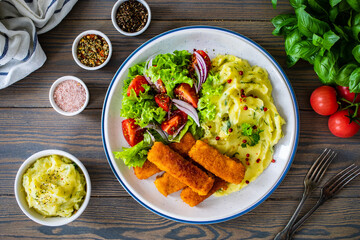 Fried fish sticks with potato puree and fresh vegetable salad on wooden table