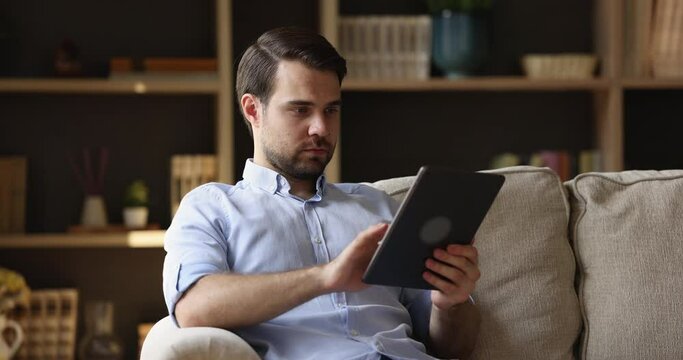 Focused millennial guy sit on couch use modern touchpad device for work study. Young man spend free time at home hold digital pad chat at social network type message on screen play virtual game in app