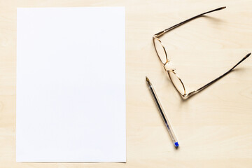 top view of eyeglasses, pen and blank sheet of white office paper on light brown wooden board