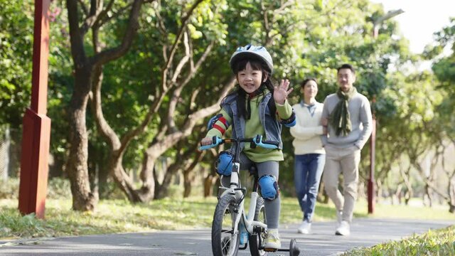 asian little girl riding bike outdoors in city park with parents walking watching in background