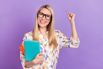 Photo of smart lucky young woman wear print shirt spectacles smiling holding book stack rising fist isolated violet color background