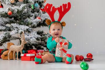 baby in a green bodysuit with red deer horns on his head is playing with gift boxes near Christmas...