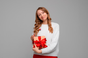 Young happy woman with blonde curly hair posing on gray background with gift box decorated with red ribbon. New Year Women's Day birthday holiday concept