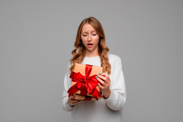 Young woman in white sweater looking inside gift box with shocked expression, secretly unpacking her gift, standing over gray background. New Year Women's Day birthday holiday concept